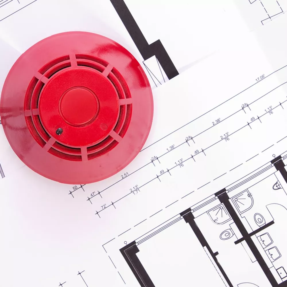 Red fire alarm device placed on architectural floor plans with technical drawings and measurements visible.