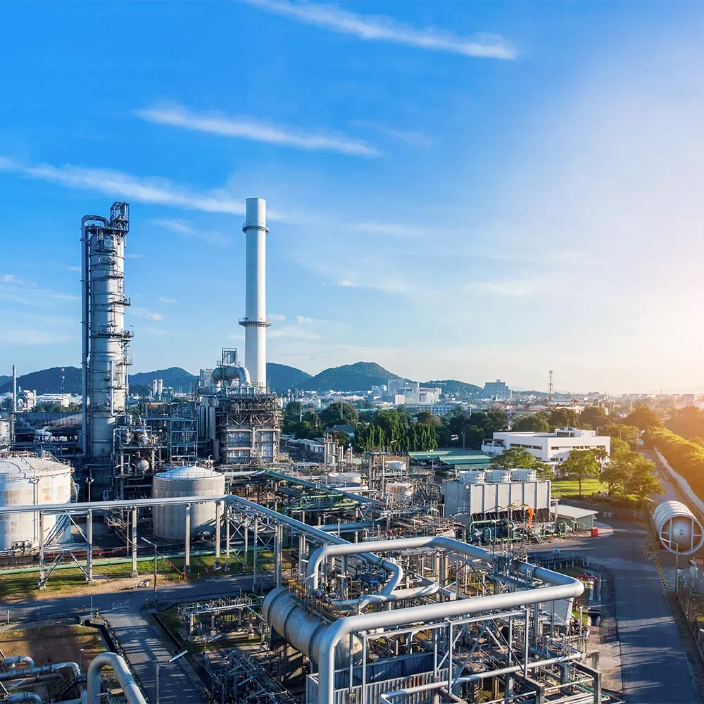 Industrial complex with pipes, storage tanks, and tall chimneys under a blue sky; mountains and a cityscape are visible in the background.