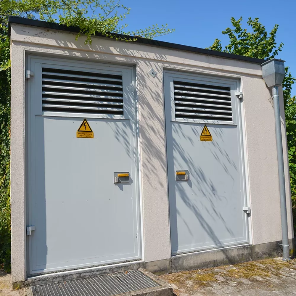Small gray building with two doors each having ventilation grilles and high voltage warning signs, surrounded by trees.