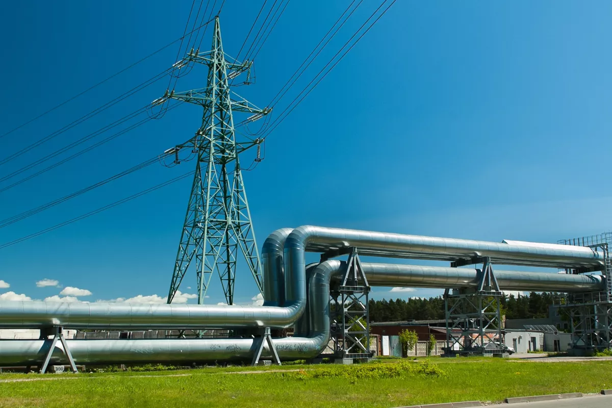 Industrial pipeline installation beneath a high-voltage transmission tower with overhead power lines under a clear blue sky.